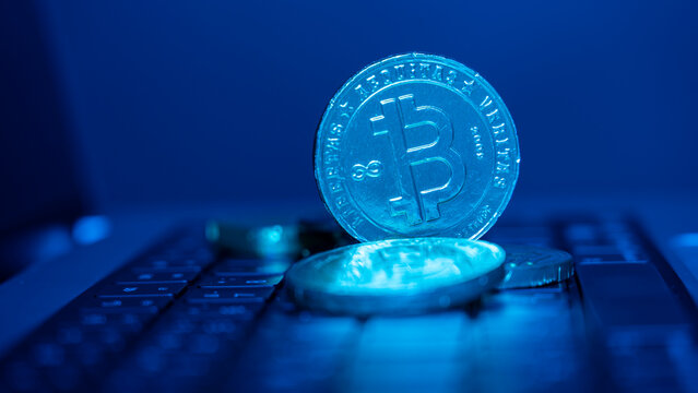 Shiny Bitcoin coin lies on a keyboard. Blue light illuminates the coin's details. The keyboard has dark keys and subtle reflections. This setup shows digital currency themes