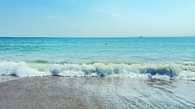 Ocean waves rolling onto a sandy beach during the day. The water is bright blue and calm, with small ripples at the shore. There are some distant boats on the horizon.