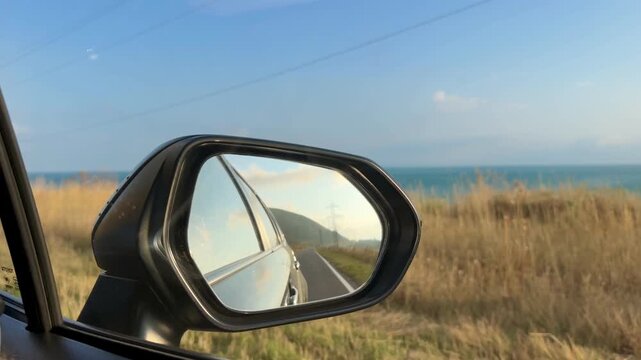 Side mirror of a car shows a coastal view with ocean waves, grassy roadside, and distant hills against a bright blue sky
