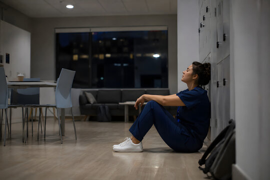 Tired young healthcare professional in scrubs sitting alone on the floor after a stressful shift, reflecting in a hospital break room and coping with workplace fatigue.