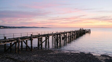 Obraz premium Old wooden pier stretching into a calm sea at sunset with a pastel sky on the horizon. Peaceful coastal scenery for travel, relaxation, retreat, and evening getaway themes. 