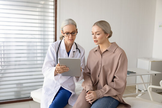 Professional female doctor showing medical information, explaining diagnose on digital tablet to woman patient during clinic appointment. Diagnosis discussion, telemedicine, healthcare consultation