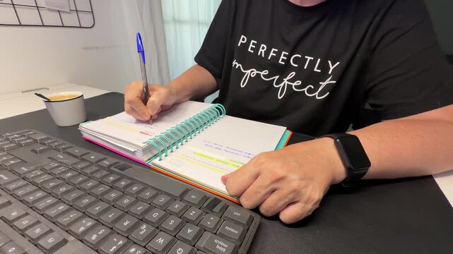 A person is sitting at a desk with a planner and pen. They are writing down tasks and notes with a keyboard nearby. This happens in a home office setting.