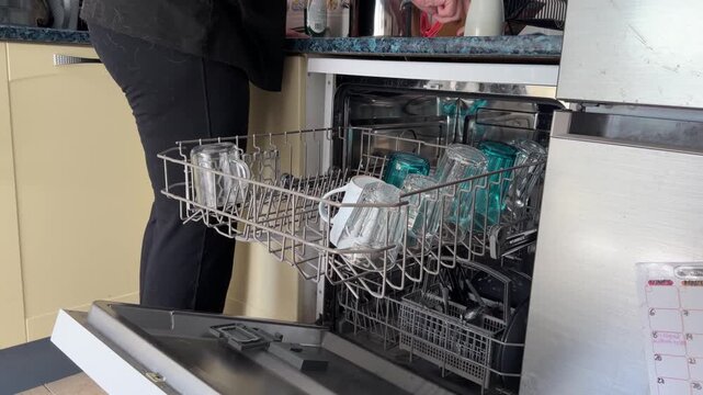 A person loads glasses into a dishwasher in a kitchen. Later, they take out clean glasses from the appliance and place them on the counter.