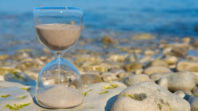 Hourglass measuring time on a rocky beach. Transparent glass hourglass with sand flowing from the top to the bottom bulb, symbolizing the passing of time, positioned on a rocky shore