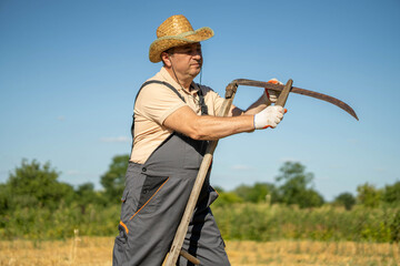A farmer stands in a field holding a scythe. He wears gloves and a straw hat. The sun shines brightly and trees are visible in the background © Віталій Б.