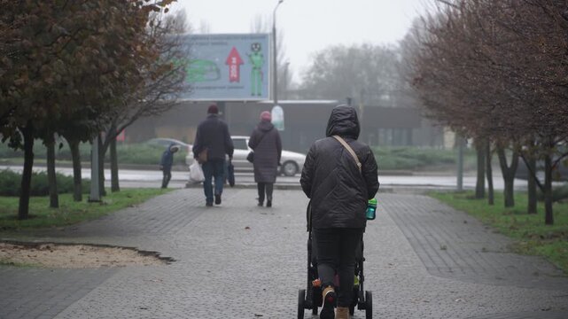 single mother, raising child alone. black jacket jeans hood over head pushes stroller sleeping autumn rain city park. cool day, filmed from side. trunk stroller full toys. background busy road with