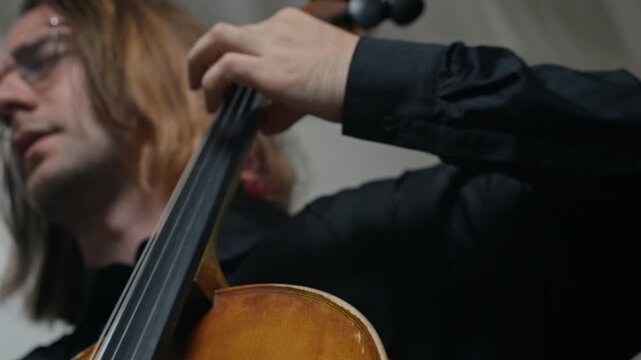 Caucasian young adult man playing cello, moving bow across strings with camera tilting up to close-up of face. Ideal for classical music promotions or arts education advertisements