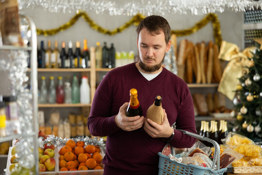 Interested man chooses wine and champagne in a supermarket with a Christmas tree in the background. The buyer chooses alcohol for the holiday