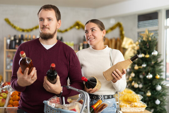 Happy young married couple choosing alcohol in grocery section of supermarket to celebrate Christmas