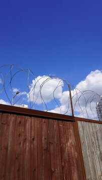 Wooden fence with razor wire against sky. Brown wooden fence panels topped with coiled barbed wire reach toward blue sky with white clouds. Security barrier creates strong vertical lines.