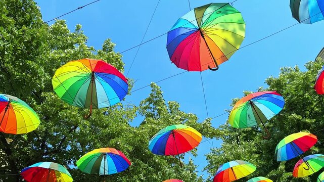 A row of bright umbrellas is suspended above a street. They are arranged in various colors, creating a festive look. The sun shines overhead on a clear blue sky.
