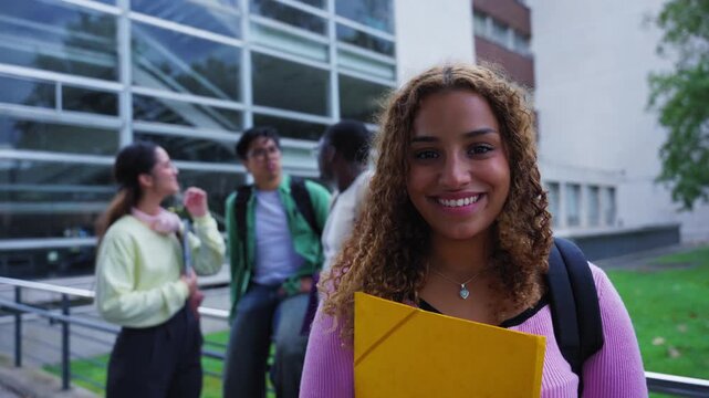 Beautiful young african american woman with afro hair smiling and looking at the camera, standing in front of the university building with other students in the background on the stairs