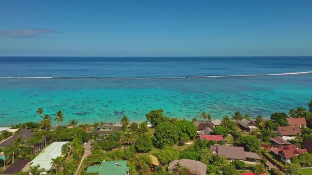 Moorea village coastal houses nestled among lush green tropical vegetation, facing vibrant turquoise lagoon, coral reef, and deep blue Pacific Ocean under a clear sky. Rural life in French Polynesia