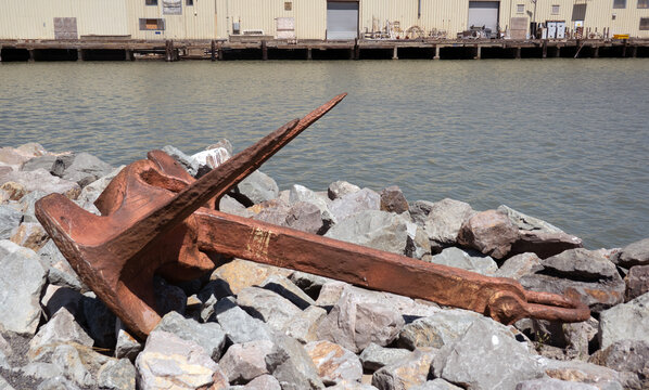 anchor-A large, rusty metal anchor rests on a rocky shoreline near a calm body of water with industrial buildings in the background.