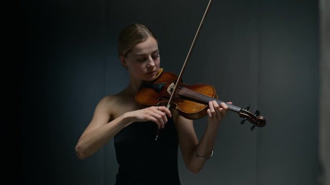 Caucasian young adult woman playing violin against dark background. Clip suitable for classical music concert promotions, artistic performance ads or music academy marketing campaigns