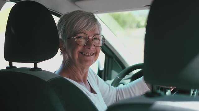 Happy senior woman with gray hair driving a car, smiling as she turns back to look at the camera. An elderly lady enjoying an active retirement, driving on a sunny day with a joyful expression