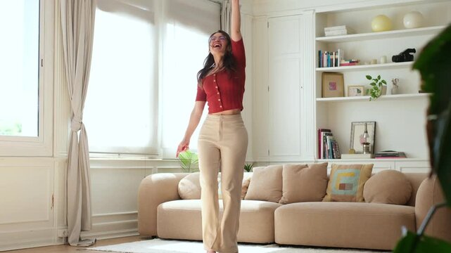 Joyful and carefree young woman dancing energetically in her modern and bright living room, celebrating freedom and happiness with expressive moves in a sunlit apartment during the day