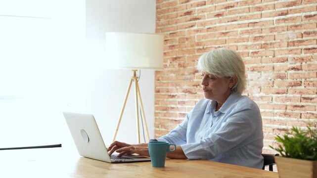 Senior woman working from home on a laptop, finishing her task and stretching her arms and back with satisfaction and relief, feeling relaxed after completing her project successfully