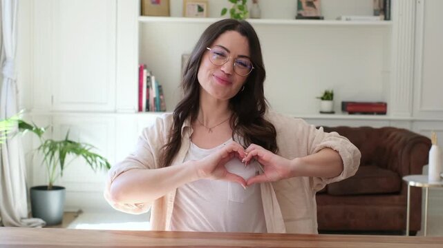 Charming young woman sitting at a desk, smiling and showing a heart sign with her fingers, expressing love, gratitude, and support while looking directly at the camera in a modern interior
