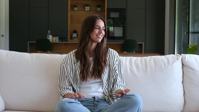 Beautiful young woman sitting comfortably on a white sofa in her modern living room, smiling and looking around as she enjoys a peaceful and relaxing moment of quiet contemplation at home