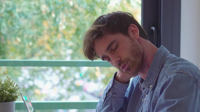 Exhausted young man experiencing stiff neck and muscle tension after long hours of remote work on his computer, feeling burnout and fatigue from a sedentary job in his home office
