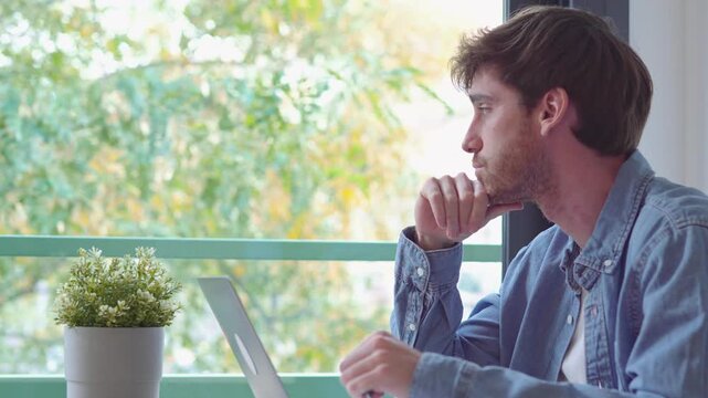 Thoughtful young man sitting at his desk with a laptop, looking out the window while thinking about a problem, pondering a solution, or feeling bored and procrastinating at home