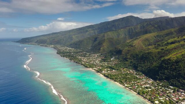 Amazing Moorea island coastline displaying its vibrant coral reef lagoon, deep blue ocean, and a coastal village nestled against the backdrop of towering green volcanic mountains under a bright sky