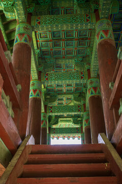 Low angle view of timber stairs and highly decorated coffered ceiling with vibrant dancheong patterns inside a traditional pavilion in South Korea.