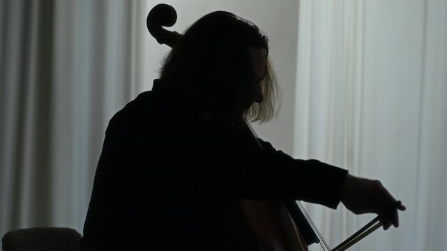 Middle aged Caucasian man playing cello in silhouette profile view near window. Video is suitable for classical music tutorials, artistic lifestyle promos, or creative documentary projects