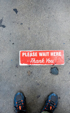 Please Wait Here-A pair of sneakers stands behind a red and white pavement sign that reads "Please Wait Here Thank You."