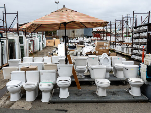 toilets-An outdoor salvage yard displays a large collection of used white toilets arranged beneath a large striped umbrella.

