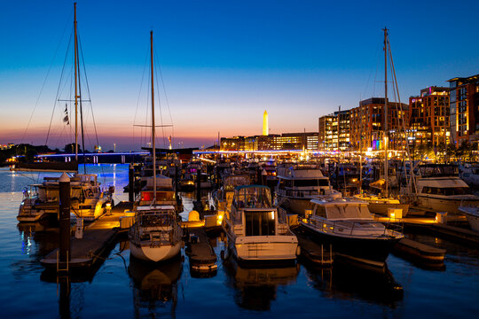 Washington DC waterfront brightens at dusk. Washington DC waterfront frames boats and yachts. City lights line Washington DC waterfront. The Washington Monument near waterfront.