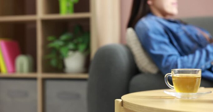 Woman with phone indoors, focus on portable audio speaker and cup of tea on wooden table. Camera moving left