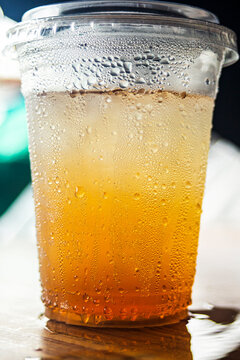 Iced amber beverage in a plastic cup with condensation on a wooden table.A refreshing eye-level shot of a cold amber-colored drink, such as iced tea, apple juice, or traditional herbal beverage