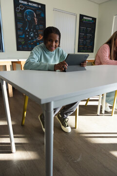 Diverse teenage classmates sitting at classroom table holding tablet, with AI posters and sunlight