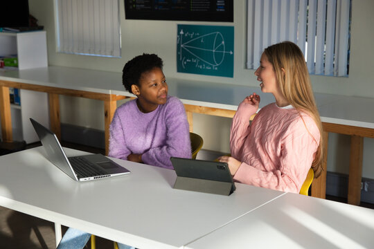 Two teenage females in lavender fuzzy and pink sweaters using laptop and tablet at classroom table