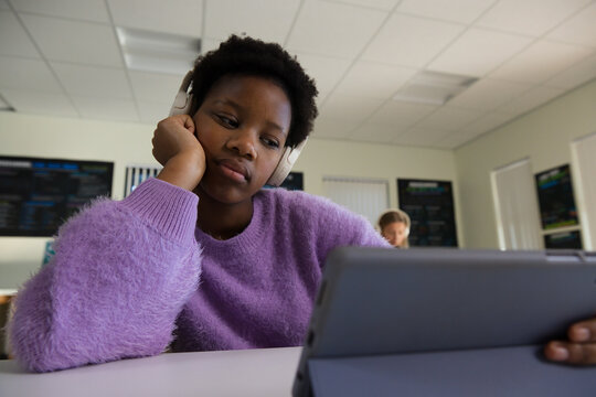 Diverse female classmates sitting in computer lab using tablet with headphones and purple sweater