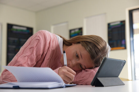 Youth girl student in pink sweater leaning at desk, writing in lined notebook using tablet folio