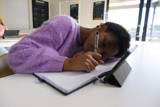 Teenage African-American student writing with pen in notebook leaning in classroom viewing tablet
