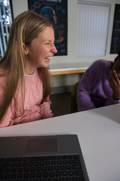 Diverse teenage female friends laughing and studying in classroom with laptop on white table