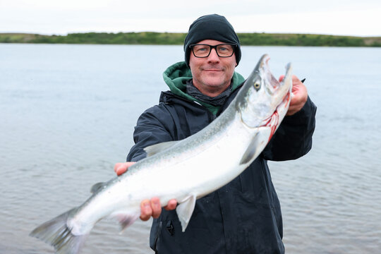 A fisherman proudly holds a large wild Coho salmon caught on the Egegik River in Alaska, displaying the silver fish against calm water and cloudy sky.