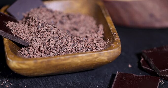 Brown dark chocolate in pieces and shavings, crushed bitter chocolate lies on a dark table next to a wooden bowl with small crumbs, plate