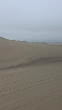 Vertical panning shot of vast sand dunes in the Ica desert near Huacachina, Peru, showing natural wind ripples and vehicle tracks under a moody overcast sky. Ideal for vertical video reels, mobile wal