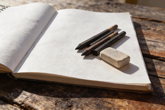 Open sketchbook with charcoal pencils and eraser on weathered wooden surface