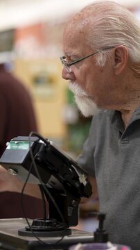 An elderly man shops in a grocery store and uses a device to examine products on a table. He is focused on his task.