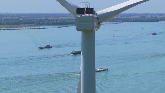 Aerial view of a wind turbine spinning its blades over a wide river. Several cargo barges navigate the waterway on a sunny day in the netherlands