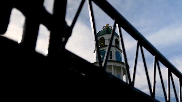 A clock tower stands tall in a town. The image shows the tower viewed through a fence. The sky is blue with some clouds. Sunlight shines on the building.