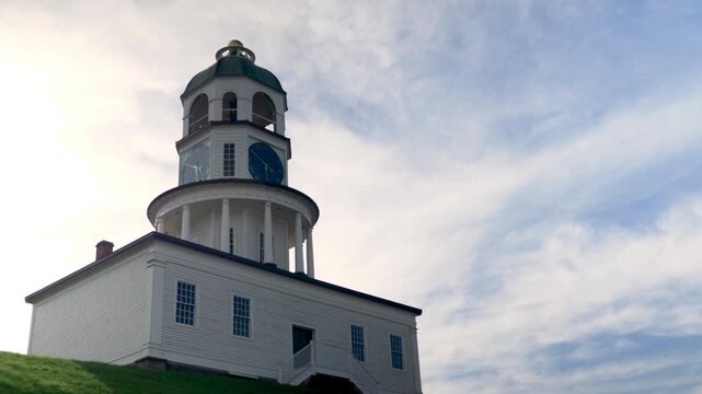 A clock tower is located on a grassy hill. The tower has a green dome and white walls. It is daytime with clouds in the sky. The area shows a coastal setting with greenery.