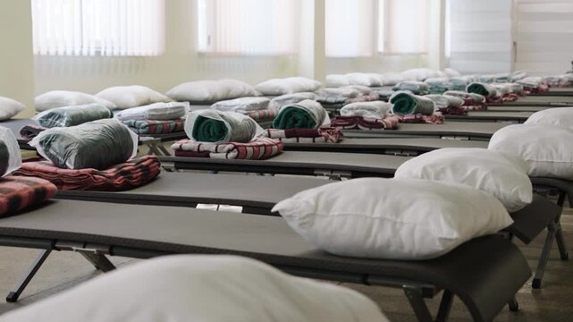In a large room, there are many cots lined up, each with neatly arranged blankets and pillows. This scene shows the preparation for guests in need during a community event.